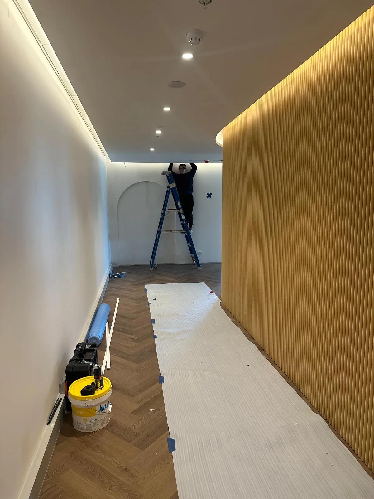 Electrician installing recessed lighting along a hallway with a wooden accent wall in a commercial space in Sydney